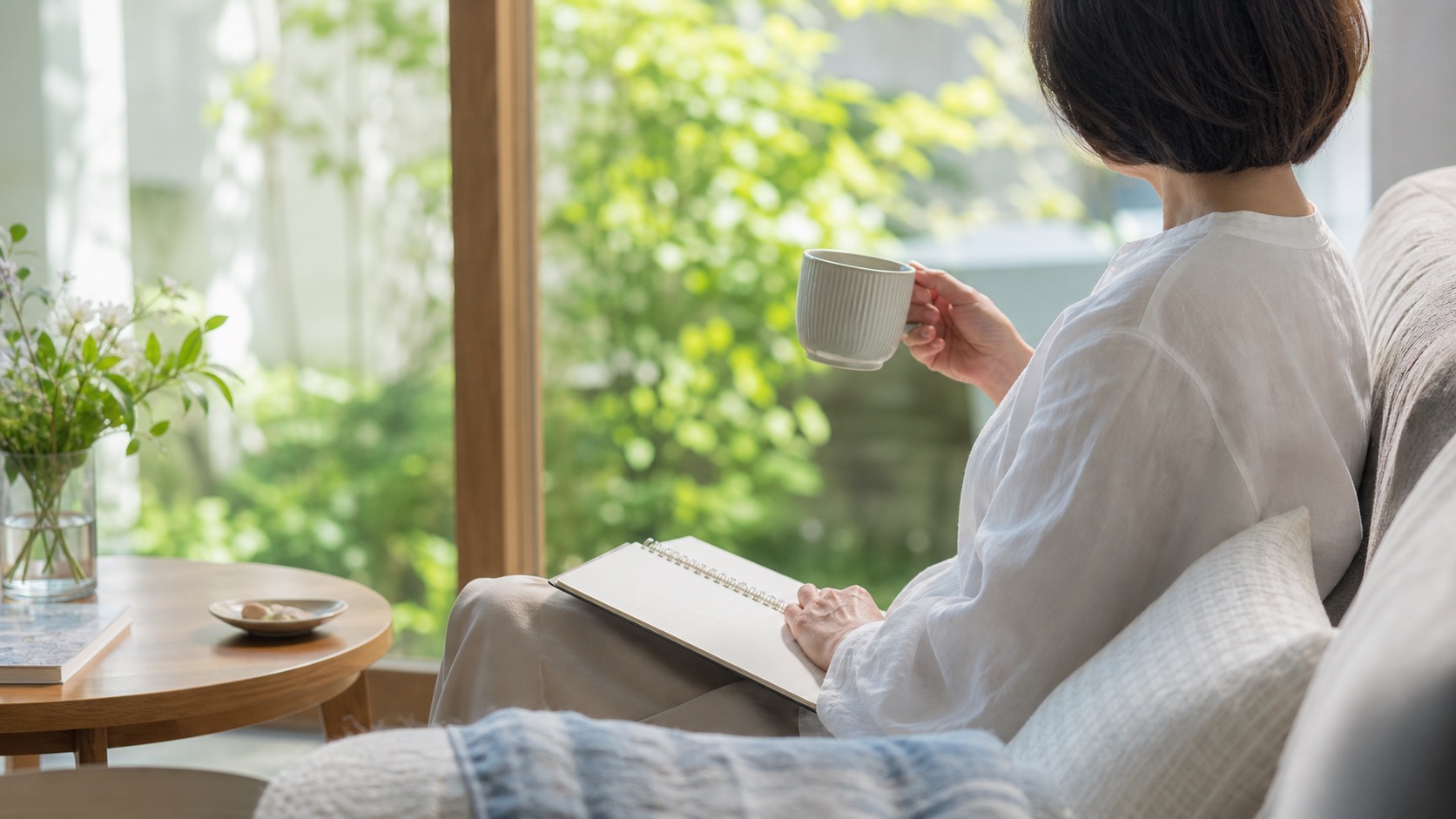 窓辺でノートとお茶を持って過ごす女性の後ろ姿